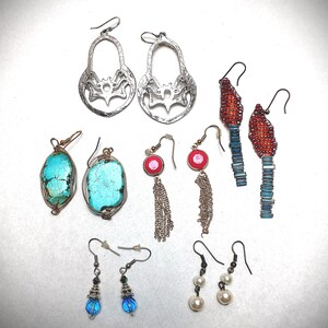 Op de afbeelding: Een verzameling handgemaakte oorbellen in verschillende stijlen. Inclusief zilverkleurige filigraan, turquoise steen gewikkeld in draad, rood-zilveren kralen, blauwe kralen en pareldruppel oorbellen. De oorbellen worden gepresenteerd op een wit oppervlak.