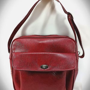 May include: A red shoulder bag with a textured surface and a flap pocket. The bag has a long, adjustable strap and a silver-colored clasp. The bag is rectangular and appears to be made of leather or a leather-like material.
