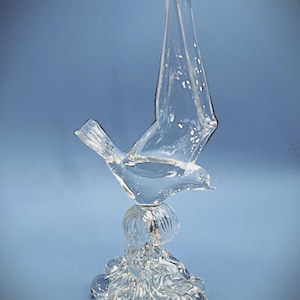 May include: A clear glass figurine of a bird in flight, perched atop a textured, wave-like glass base. The bird is depicted with wings extended upward, and the entire piece is crafted from transparent, colorless glass with smooth surfaces.