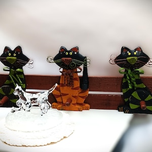 Puede incluir: Tres ganchos de metal con forma de gato negro y verde con detalles naranja y marrón están montados en un estante de madera. Los ganchos están diseñados para parecer gatos sentados en un banco. Una pequeña figura de gato de vidrio transparente está delante del estante.