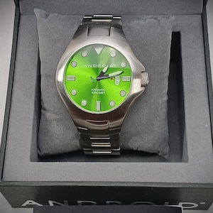 May include: A stainless steel Android automatic watch with a bright green face. The watch has a silver band and a date display at the 3 o'clock position. The watch is presented in a gray box with a gray pillow.