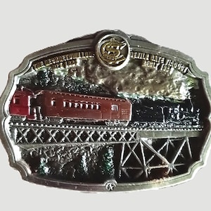 May include: An oval metal belt buckle featuring a relief scene of a train crossing a trestle bridge. The text reads THE GEORGE P.I.V.A. LOG. BUILT 1887 and DEVILS GATE VIADUCT BUILT 1881. A gold-toned circular emblem with the letters CS is at the top center.