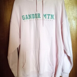 Puede incluir: Una sudadera con capucha rosa con el texto "Gander MTN. We Live Outdoors" en verde.