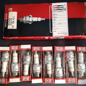 Può includere: Una scatola di otto candele Champion RJ12YC con resistenza, etichettata "STK NO. 14" e "Nothing Sparks Like A Champion". La scatola è rossa con testo nero e un'etichetta bianca. Le candele sono confezionate singolarmente in scatole di plastica trasparente con etichette rosse e bianche.