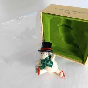 Peut inclure: Une décoration de Noël en forme de bonhomme de neige blanc portant un chapeau haut de forme noir, une écharpe rouge et une écharpe à carreaux verts. L'ornement est dans une boîte avec le texte "Hallmark Tree-Trimmer Collection".