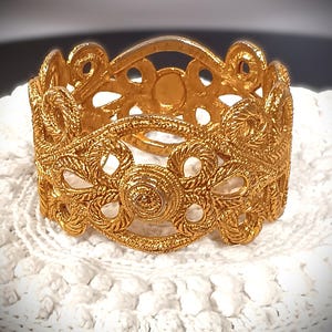 May include: A gold-toned metal bracelet with a delicate, intricate design. The bracelet is made of a series of interconnected circles and swirls.