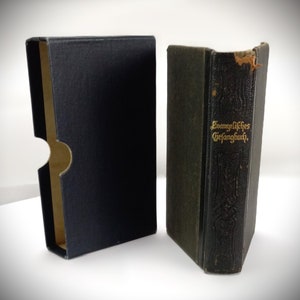 May include: A black book with gold lettering on the spine that reads "Evangelisches Gesangbuch". The book is in a black box.