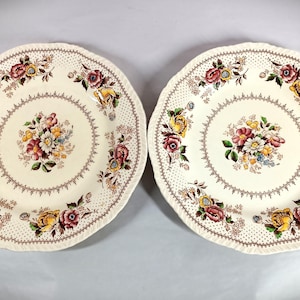 May include: Two cream-colored, scalloped-edge plates with floral designs. Each plate features a central bouquet of flowers in shades of red, yellow, and white, surrounded by a dotted border and floral accents. The plates are likely for serving.