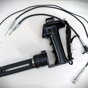 May include: Black camera grip with a black handle and a black and silver rail system. The grip has a black strap and the text "S/1172" printed on it. The grip is connected to a black cable with a silver connector.