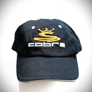 Könnte beinhalten: Eine schwarze Baseballkappe mit einer gestickten goldenen Krone und einem stilisierten goldenen Schlangenlogo über dem weißen Schriftzug "cobra" auf der Vorderseite. Die Kappe hat einen gebogenen Schirm mit einem dünnen weißen Rand und ein sechsteiliges Design.