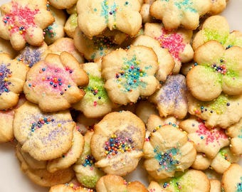 May include: A collection of golden-brown, flower-shaped butter cookies topped with colorful sugar sprinkles in shades of pink, blue, green, and purple. The cookies are arranged in a dense pile, showcasing their scalloped edges and textures.
