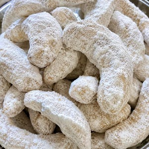 Può includere: Una collezione di biscotti a forma di mezzaluna disposti in un mucchio. Ogni biscotto ha una consistenza dorata e cotta ed è generosamente ricoperto da un sottile strato di zucchero a velo bianco, creando un aspetto uniforme e innevato.