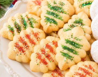 May include: A collection of golden-brown, tree-shaped butter cookies arranged on a white scalloped platter. The cookies are decorated with horizontal stripes of red and green sugar sprinkles, alongside several round, white powdered sugar cookies.