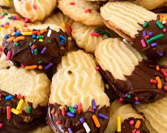 May include: A pile of light-colored, ridged butter cookies, many of which are partially dipped in dark chocolate and topped with colorful, multi-shaped sprinkles. The cookies have a distinct piped texture and are arranged in a dense, overlapping cluster.