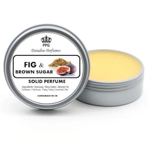 May include: A silver tin with a white label that reads "PPG Paradise Perfumes Fig & Brown Sugar Solid Perfume". The label also includes a crown logo, a drawing of a fig, and the ingredients: beeswax, shea butter, almond oil, perfume, patchouli, and ylang ylang essential oils. The tin is filled with a yellow solid perfume.