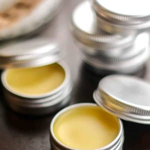 May include: Close-up of four small silver tins with yellow balm inside. The tins are stacked on top of each other. The balm is a light yellow color and has a smooth texture.