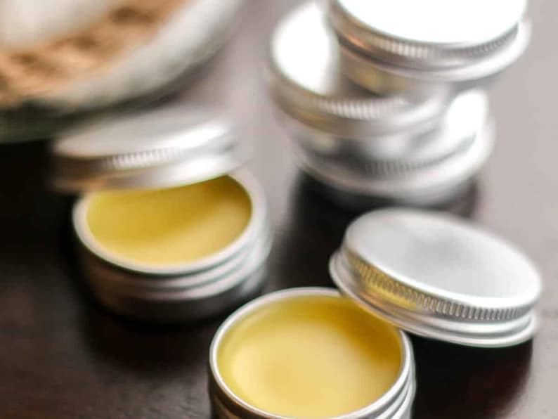 May include: Close-up of four small silver tins with yellow balm inside. The tins are stacked on top of each other. The balm is a light yellow color and has a smooth texture.