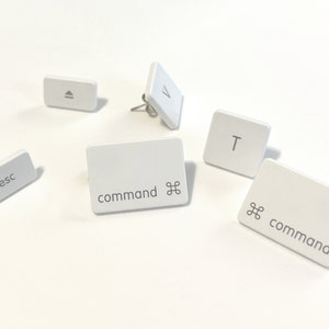 Puede incluir: Cinco pendientes de plástico blanco en forma de teclas con postes plateados. Las teclas presentan las letras "T", "V", "esc" y las palabras "command" con el símbolo de comando de Apple.