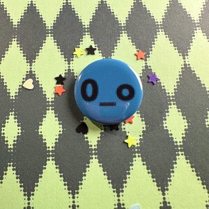 May include: A circular blue pinback button featuring a black pixelated face with two circular eyes and a straight horizontal line for a mouth, surrounded by small scattered star and heart-shaped confetti pieces.