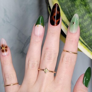 May include: Close-up of a hand with green, brown, and white nail polish. The nails are decorated with a flower, a star, and a leopard print. The hand is wearing several gold rings.