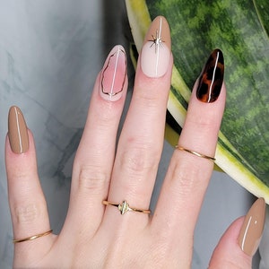 May include: A hand with almond-shaped nails painted in various designs. The nails are painted in shades of brown, pink, and white. Some nails have geometric designs, while others have a leopard print. The hand is wearing several gold rings.