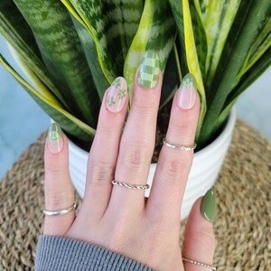 May include: A hand with green and white checkered nail polish and green flower nail art. The hand is wearing several silver rings. The hand is in front of a green plant.
