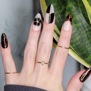May include: Close-up of a hand with almond-shaped nails painted with a black and white geometric design, a brown and gold swirl design, and a black flower design. The hand is wearing several gold rings.