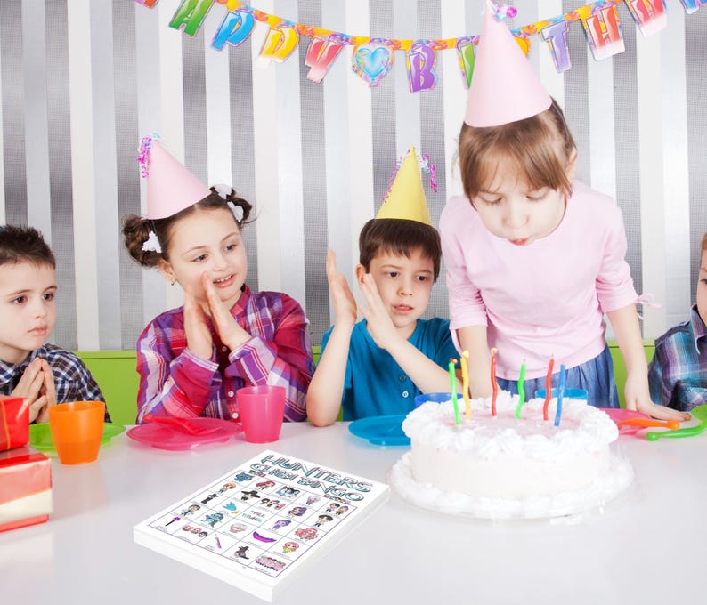 May include: A group of children at a birthday party. A white cake with lit candles sits on a table. A game board with the text "Hunters Queen Bingo" is in the foreground. Children wear party hats and celebrate.