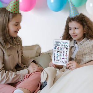 May include: Two young people wearing party hats are seated, holding a bingo card. The card, titled "Hunters Chibi Bingo," features various cartoon illustrations. Colorful balloons decorate the background, creating a festive atmosphere.