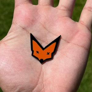 May include: A hand holding a small orange fox pin with black outlines and x's for eyes.