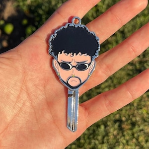 May include: A silver key chain featuring a cartoon portrait of a person with black hair, wearing sunglasses and a nose ring. The person has a light brown complexion and a serious expression.