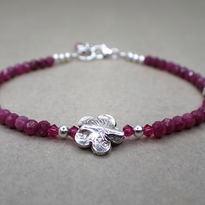 May include: A delicate bracelet featuring small, faceted, deep pink beads. A silver-colored flower charm is the focal point, with additional pink crystals and silver beads. The bracelet is secured with a silver clasp.