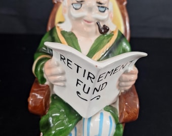 May include: Ceramic figurine of an elderly man in a green robe, seated in a brown armchair. He is reading a newspaper with the words "RETIREMENT FUND" printed on it. The figurine has a pipe in its mouth and is wearing blue and white striped pants.