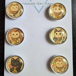 May include: A set of six handmade glass magnets featuring illustrations of the Japanese Maneki Neko cat. The cats are in various poses and colors, including tan and black, with floral backgrounds. The card reads "Cabochon Design Studio."