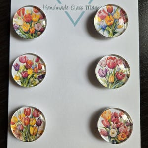 May include: Six round glass magnets with floral designs featuring tulips in various colors, including red, yellow, and purple. The magnets are arranged on a white card with the text "Cabochon Design Studio" and "Handmade Glass Magnets".
