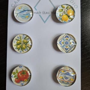 May include: Six circular glass cabochons arranged on a white display card labeled Cabochon Design Studio. The designs feature a blue fish, yellow lemons, green olives, a blue and white geometric tile pattern, red tomatoes, and a fish on a plate.
