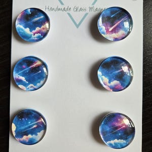 May include: Six handmade glass magnets with a celestial design. Each round magnet features a vibrant image of a night sky with shooting stars, clouds, and a mix of blue, purple, and pink hues. The card reads "Cabochon Design Studio".