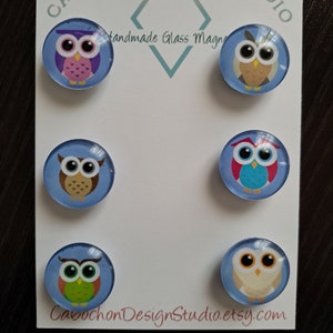 May include: Six round glass magnets with different owl designs. The owls are on a blue background. The magnets are handmade and say "Cabochon Design Studio.etsy.com".