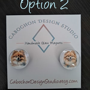 May include: Two round glass magnets with a watercolor image of a Pomeranian dog on each. The magnets are on a white card with the text "Cabochon Design Studio" and "Handmade Glass Magnets".