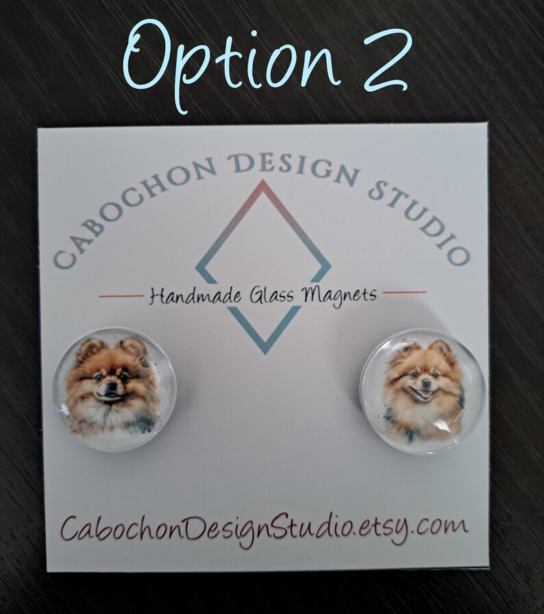 May include: Two round glass magnets with a watercolor image of a Pomeranian dog on each. The magnets are on a white card with the text "Cabochon Design Studio" and "Handmade Glass Magnets".