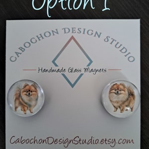 May include: Two round glass magnets featuring a brown and white Pomeranian dog. The magnets are on a white card with the text "Cabochon Design Studio" and "Handmade Glass Magnets".