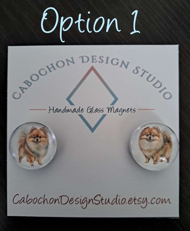 May include: Two round glass magnets featuring a brown and white Pomeranian dog. The magnets are on a white card with the text "Cabochon Design Studio" and "Handmade Glass Magnets".