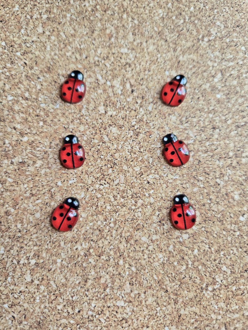 May include: Six small, red ladybug-shaped embellishments with black spots and black heads are arranged on a corkboard surface. The ladybugs are evenly spaced and appear to be made of a glossy material.