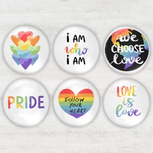 May include: Six white round buttons with colorful designs and text. The buttons feature rainbow hearts, the phrases "I AM who I AM", "we CHOOSE love", "PRIDE", "Follow YOUR HEART", and "LOVE is love".