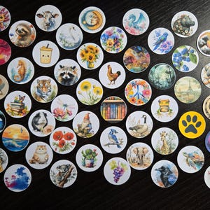May include: A collection of round, decorative buttons featuring various illustrations. Designs include animals like a sloth, cow, and tiger, along with nature scenes, food, and other whimsical images. The buttons are arranged on a dark surface.