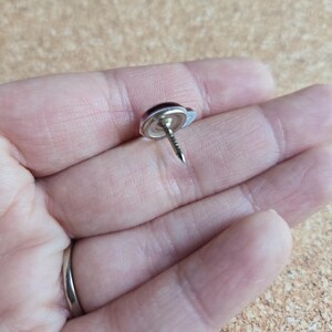 Puede incluir: Una chincheta plateada con una cabeza redondeada de color rojo oscuro. La chincheta tiene una cabeza peque&ntilde;a y circular y un alfiler afilado y puntiagudo. La chincheta est&aacute; dise&ntilde;ada para asegurar papel u otros materiales a una superficie.