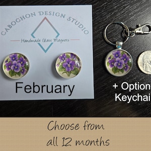 May include: Two round glass magnets and one matching keychain featuring purple violet flower illustrations on a white display card labeled February. A US quarter coin is placed next to the keychain for scale. The text reads Cabochon Design Studio.