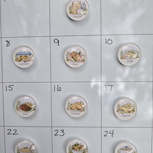 May include: A set of round, clear glass magnets featuring colorful illustrations of various food items and meals, each labeled with text including Takeout, Leftovers, Pork, Fish, Beef, Chicken, Tacos, Pasta, Meatless, and Slow Cooker.