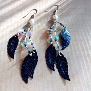 Può includere: Un paio di orecchini pendenti con ciondoli a forma di foglia nera, paillettes iridescenti e perline bianche.
