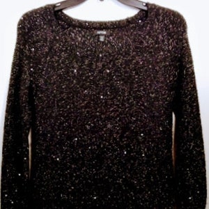 Può includere: Un maglione nero scintillante con scollo rotondo. Il maglione è realizzato in un materiale morbido e confortevole e ha un leggero luccichio.